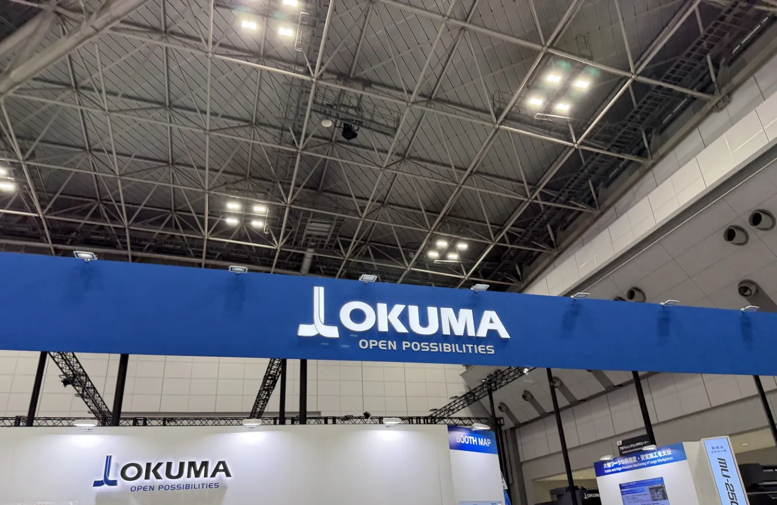 JIMTOF2026の会場。高い天井の下に、青地に白い文字で「OKUMA OPEN POSSIBILITIES」と書かれたオークマのブースの看板。